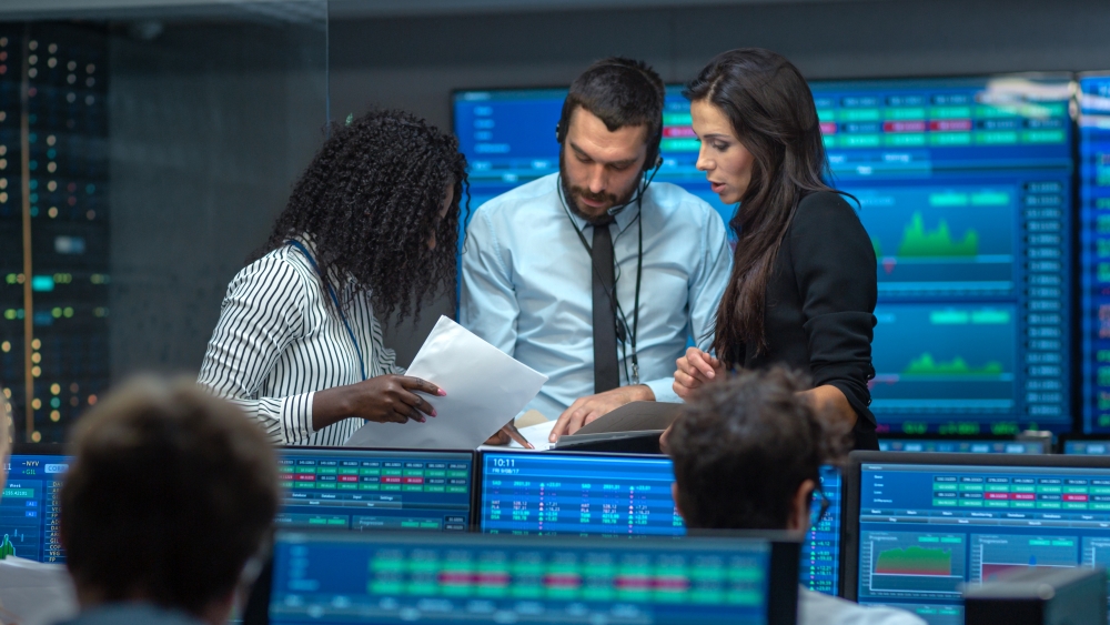 a small group of people talking on a trading floor with computer screens and large screens showing numbers surrounding them